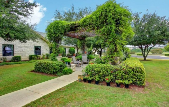 A beautifully landscaped garden featuring a pergola covered with lush greenery and surrounded by well-maintained shrubs and a paved pathway.