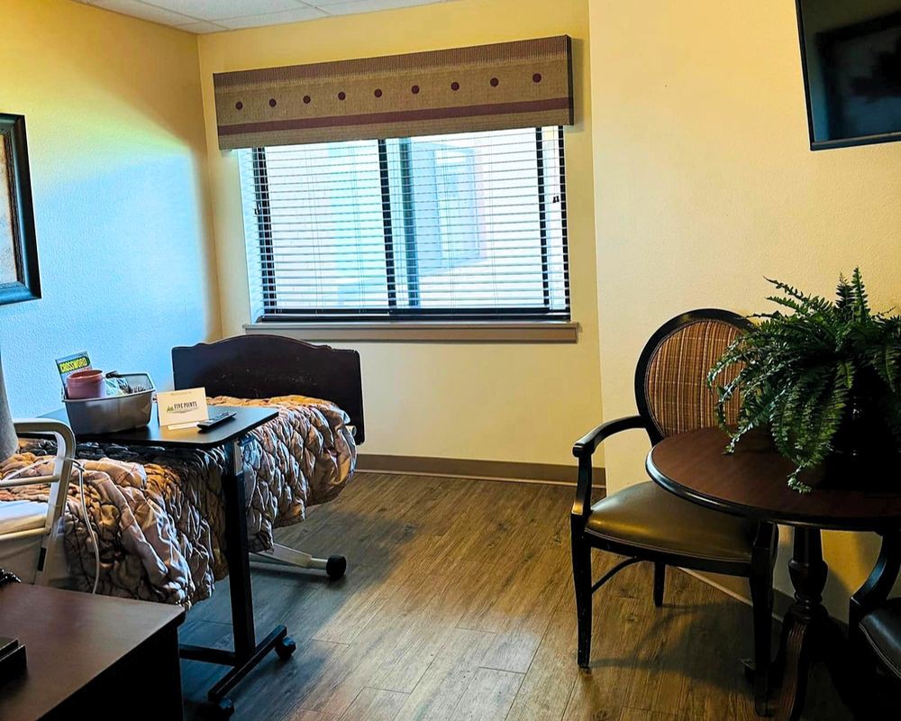 A tidy hospital room with a bed near the window, a small table with chairs, and a lamp on the nightstand.