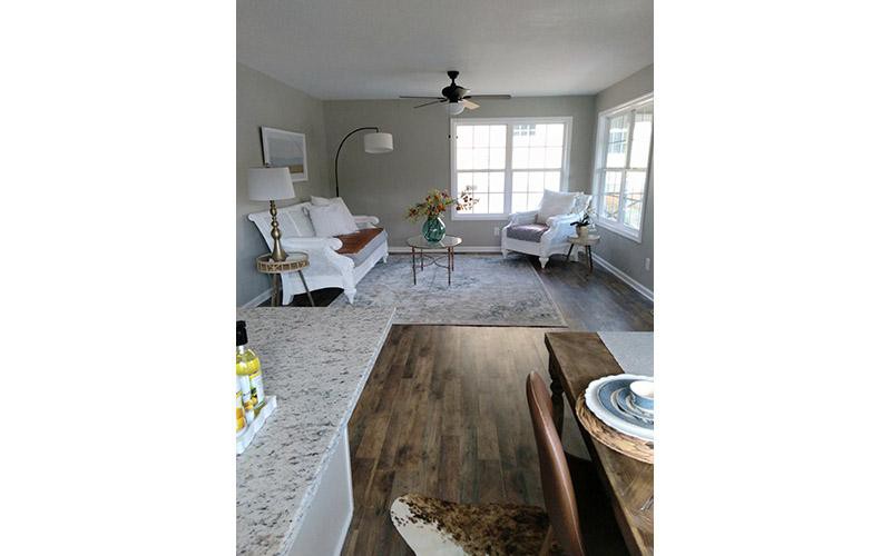 A modern living room with a neutral color palette featuring a couch, armchair, coffee table, and hardwood flooring, illuminated by natural light from large windows.