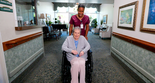 A caregiver assists an elderly woman in a wheelchair at a healthcare facility.