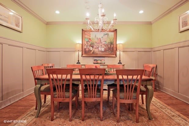 A traditional dining room with a large table and chairs, illuminated by an elegant chandelier and adorned with a classical painting.