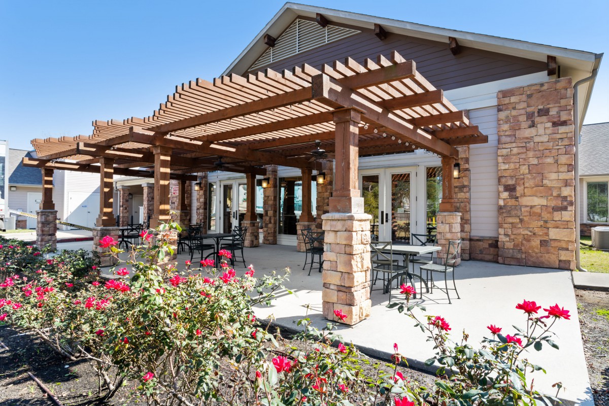 A sunny patio with a wooden pergola and outdoor seating, surrounded by blooming flowers.