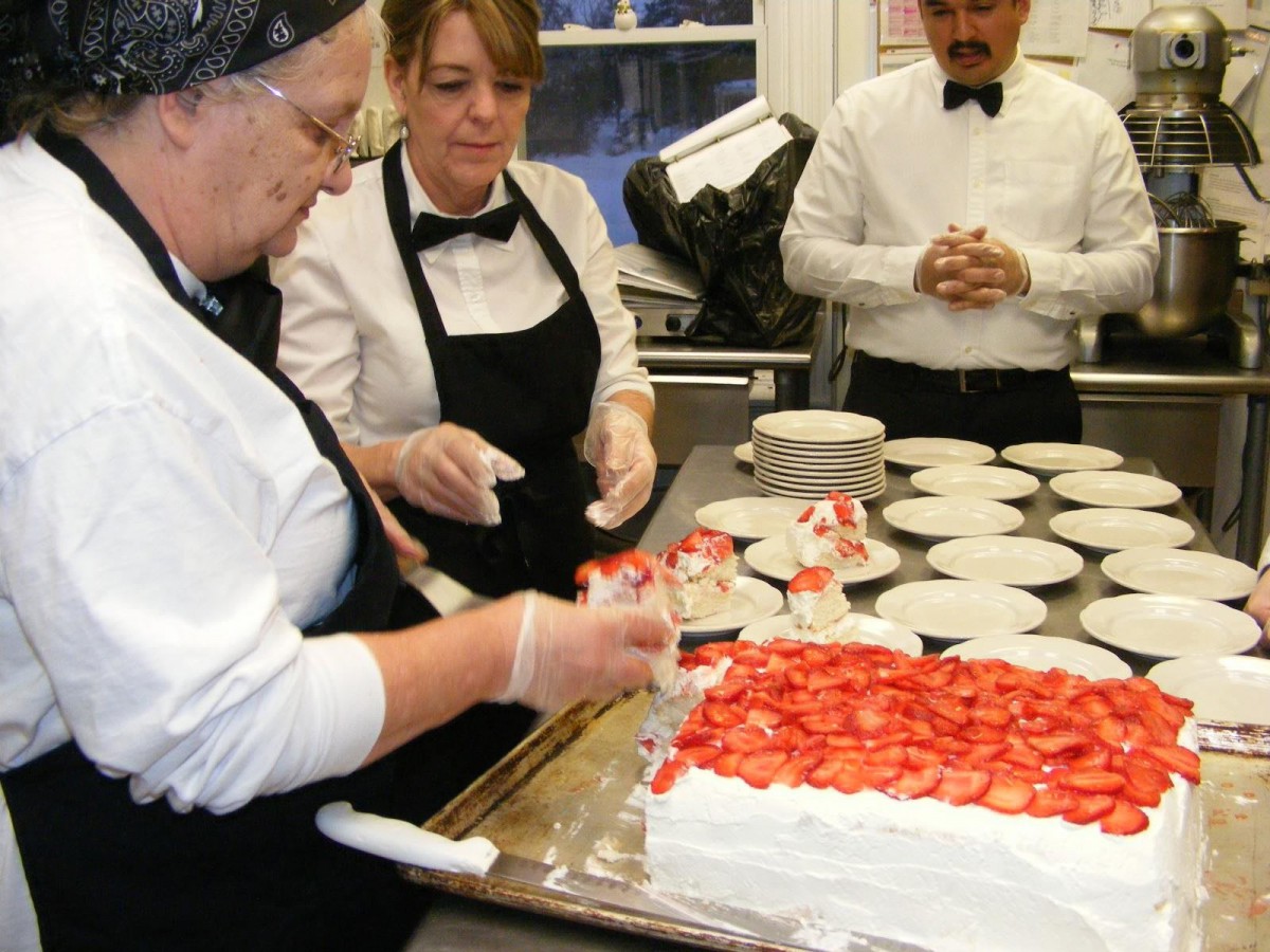 Chefs preparing a dessert topped with strawberries in a kitchen.