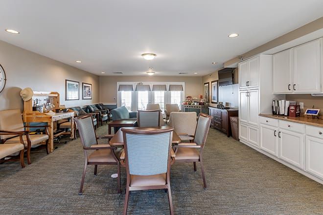 A spacious living room with chairs and tables arranged around a central area, featuring large windows and warm decor.