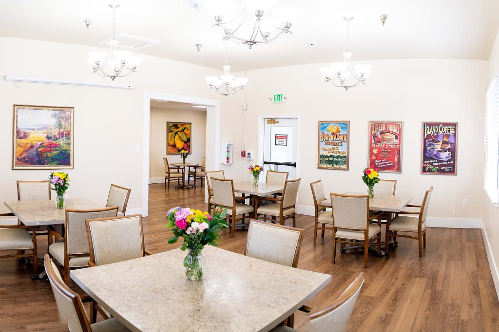 A well-lit dining area with several tables and chairs, decorated with flowers and artwork on the walls.