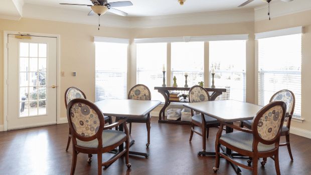 A bright dining room with several tables and chairs placed on a wooden floor, illuminated by natural light from large windows.