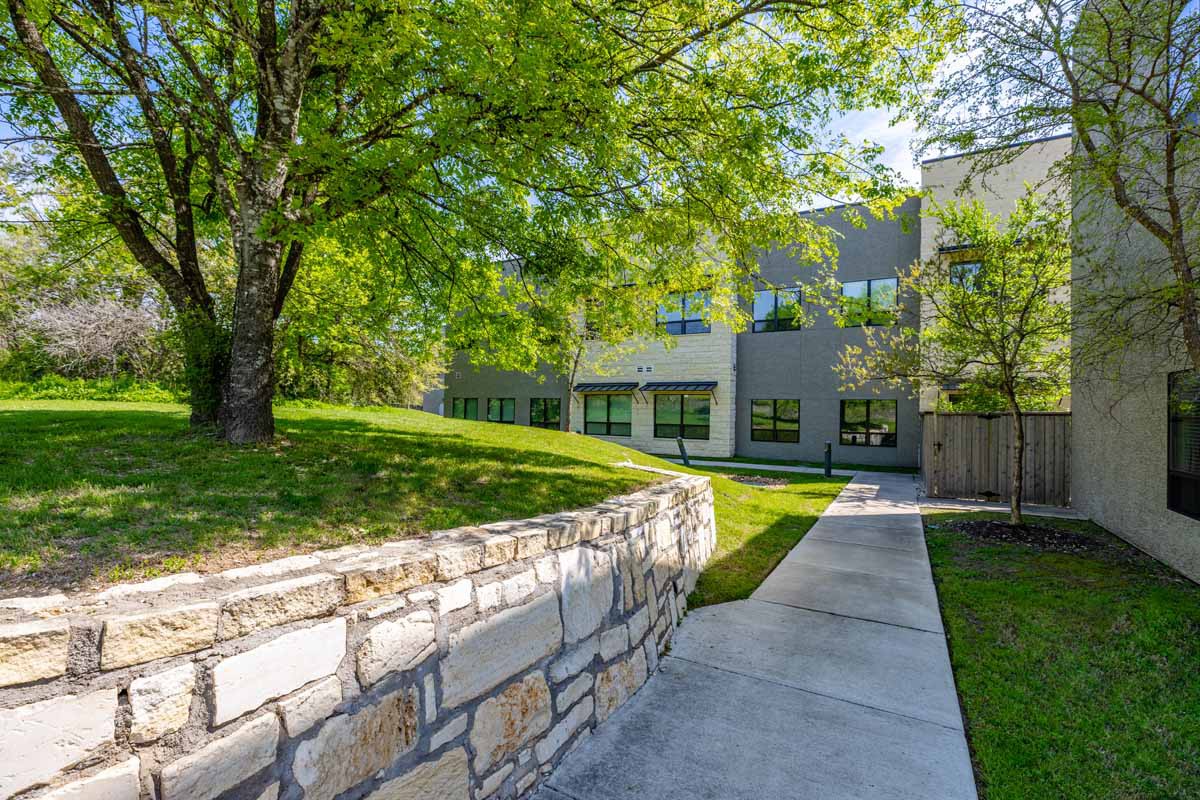 A serene outdoor scene featuring a pathway alongside a stone wall, with a modern building in the background surrounded by lush green trees and grass.