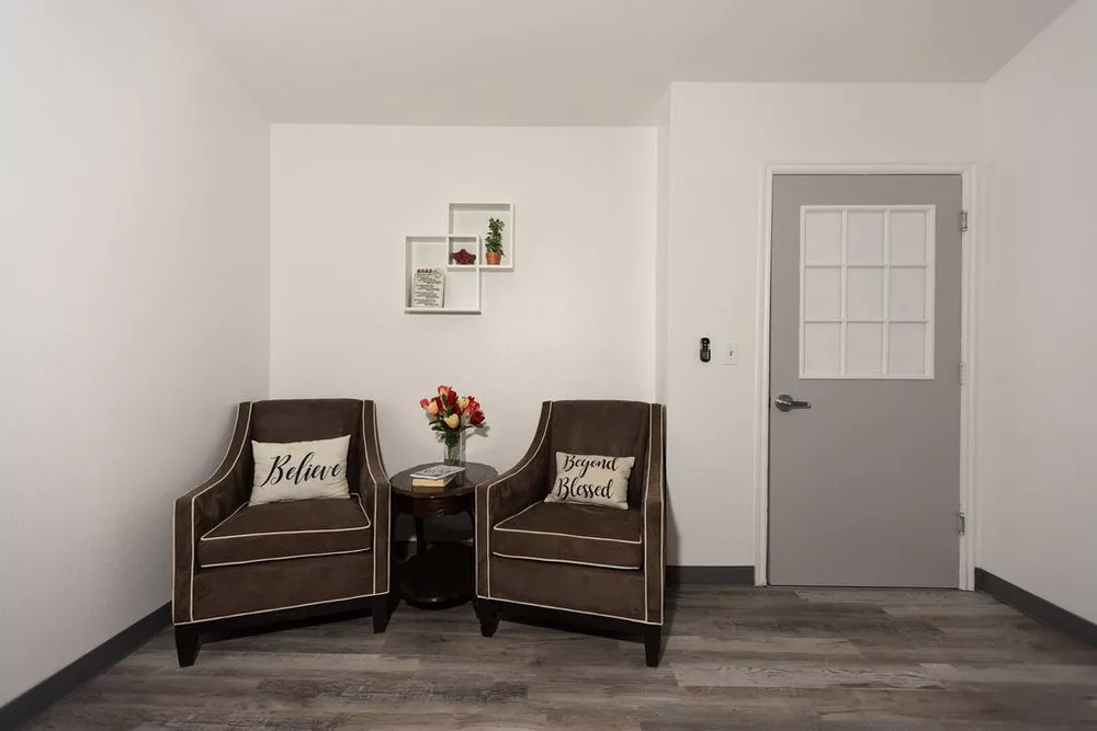 A minimalist living room corner featuring two brown armchairs with decorative pillows, a small round table with a vase of flowers, and a wall-mounted shelf.