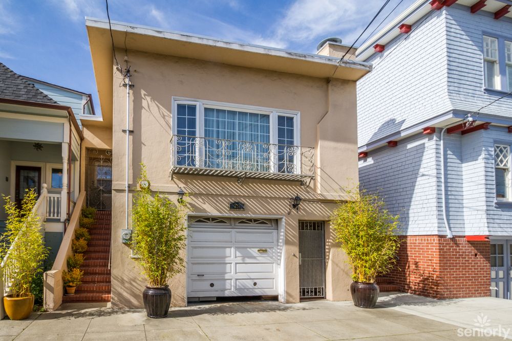 A two-story urban house with a garage and balcony, featuring potted plants on either side of the entrance.