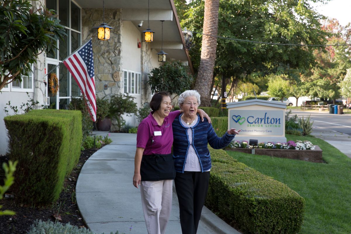 A caregiver and an elderly woman walk arm in arm outside a senior living community.