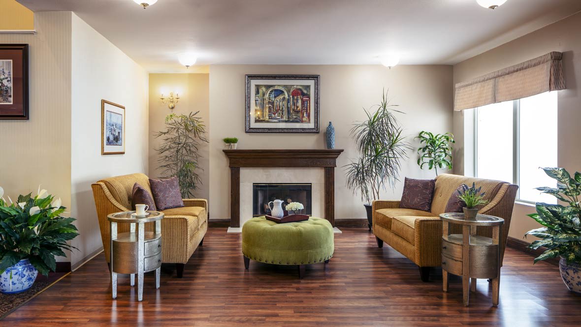 A cozy living room with a fireplace, featuring comfortable sofas, artwork on the wall, and lush green plants.