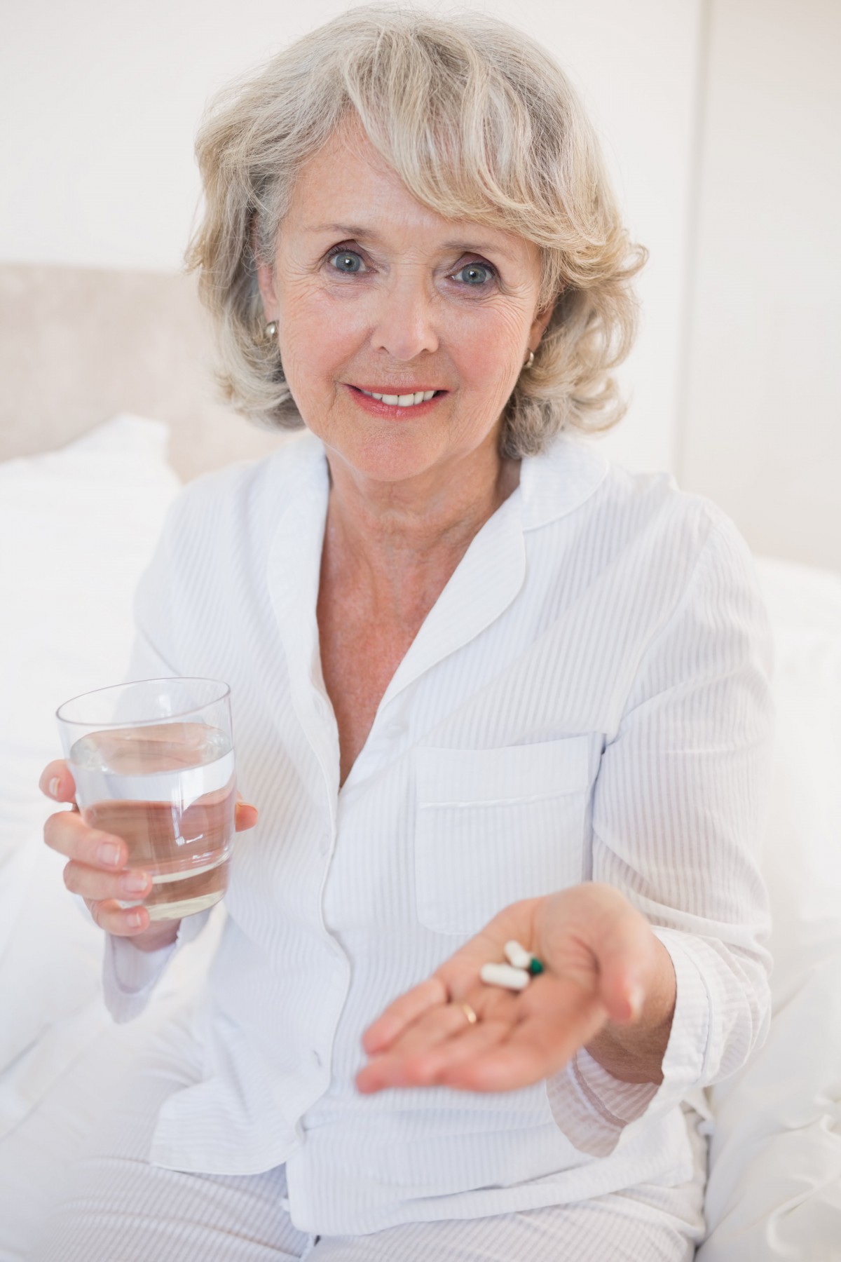 An elderly woman smiling while holding a glass of water and a handful of pills.