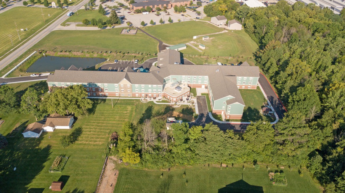 Aerial view of a large building complex surrounded by greenery and trees, with a parking lot in the background.