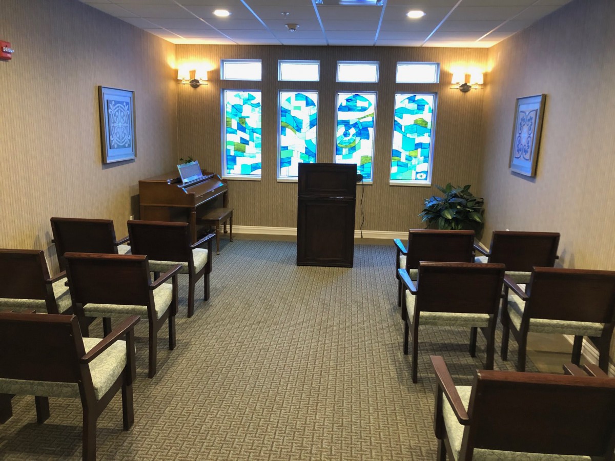 A small chapel interior featuring rows of seating, a pulpit at the front, and stained glass windows.