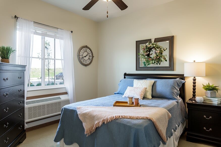 A cozy bedroom with a neatly made bed, decorative pillows, and a wall clock, complemented by a large window providing natural light.