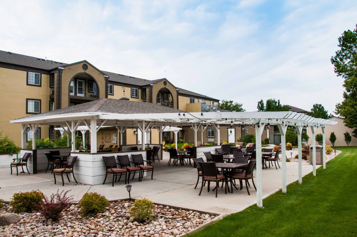 An outdoor patio area with tables and chairs under a pergola, set against a two-story building and landscaped garden.