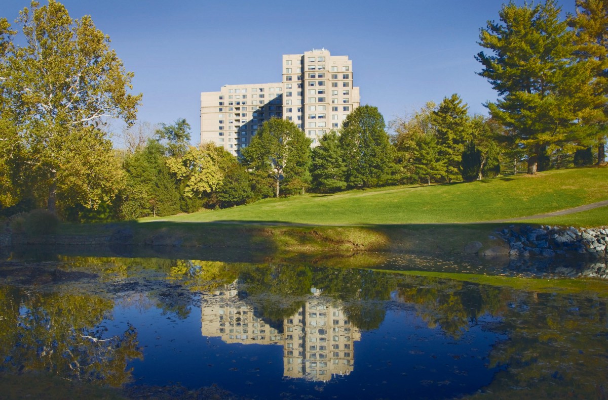 A tall building surrounded by trees reflected in a calm lake under a clear blue sky.