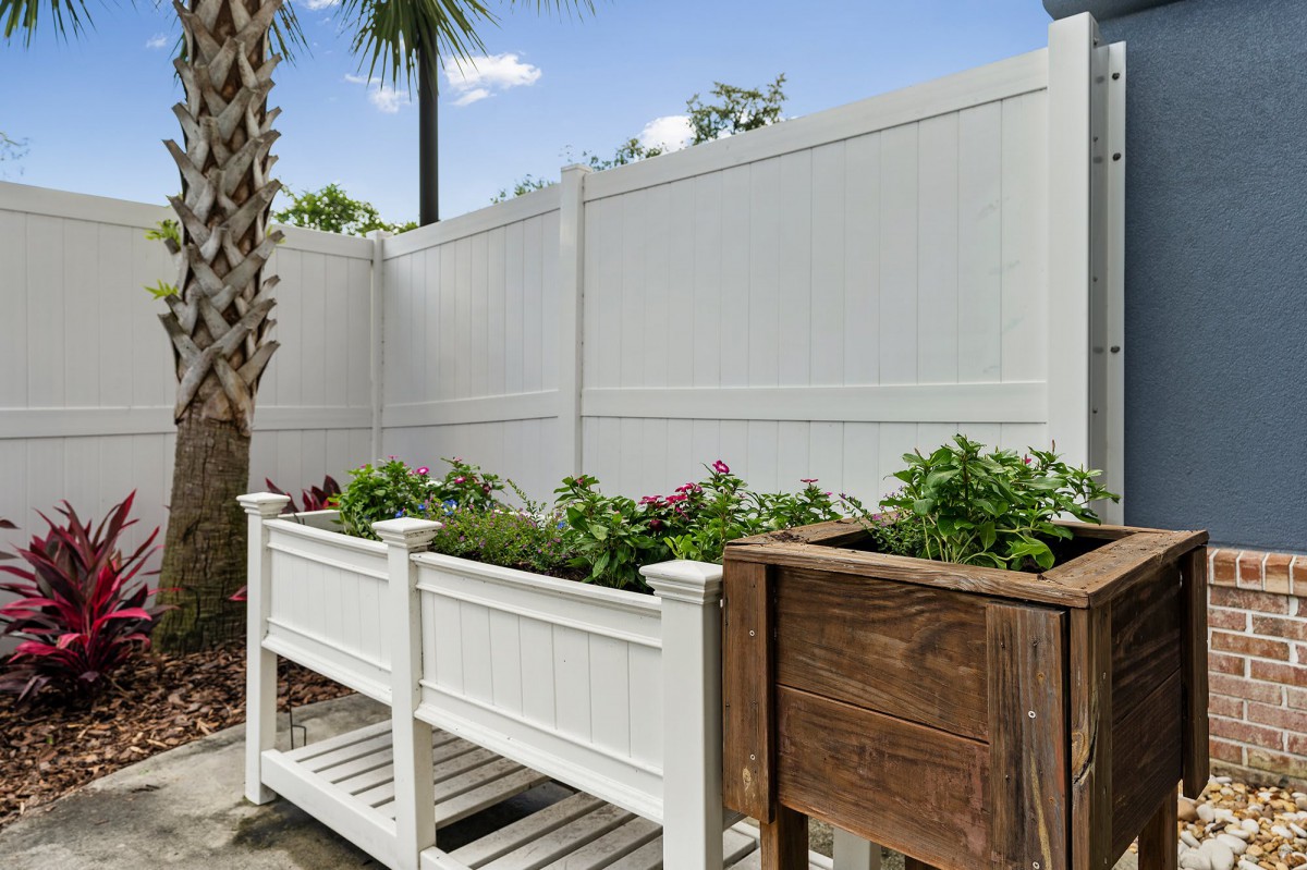 Raised planters with lush greenery in a backyard setting, bordered by a tall white fence.