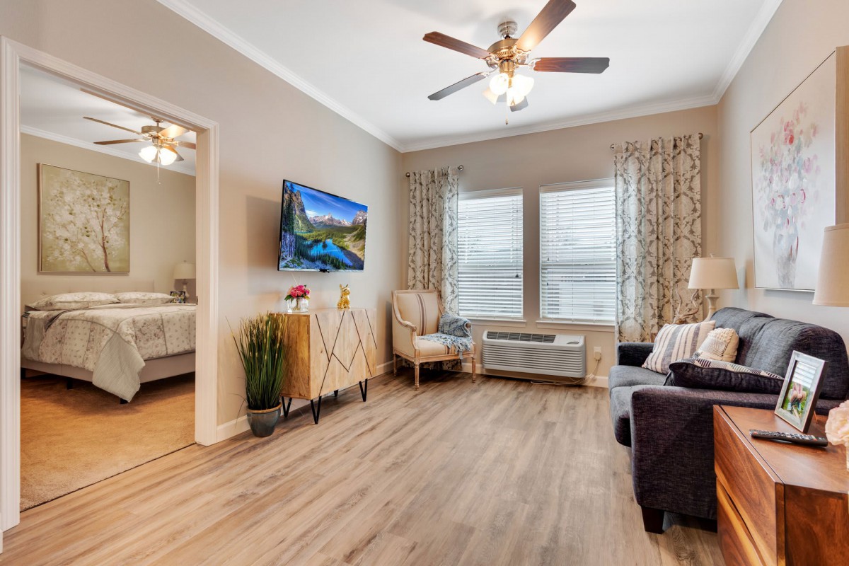 A modern living room with wood flooring and stylish decor, featuring a TV, armchair, sofa, and large windows with curtains.