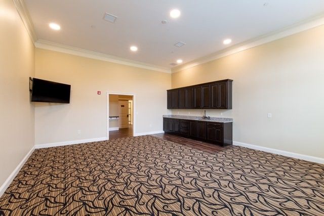 A spacious conference room with patterned carpeting, wall cabinets, and a mounted TV.