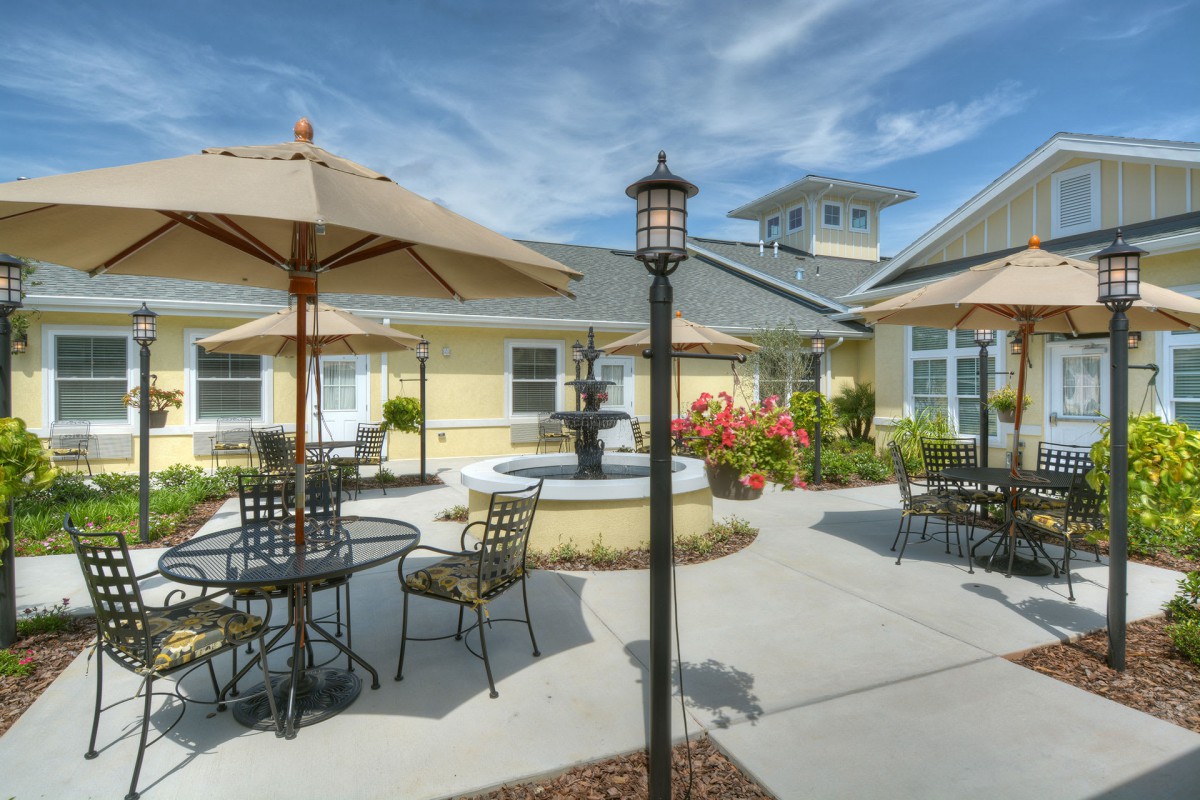 A sunny outdoor patio area featuring tables and chairs with umbrellas, a central fountain, and well-maintained landscaping.