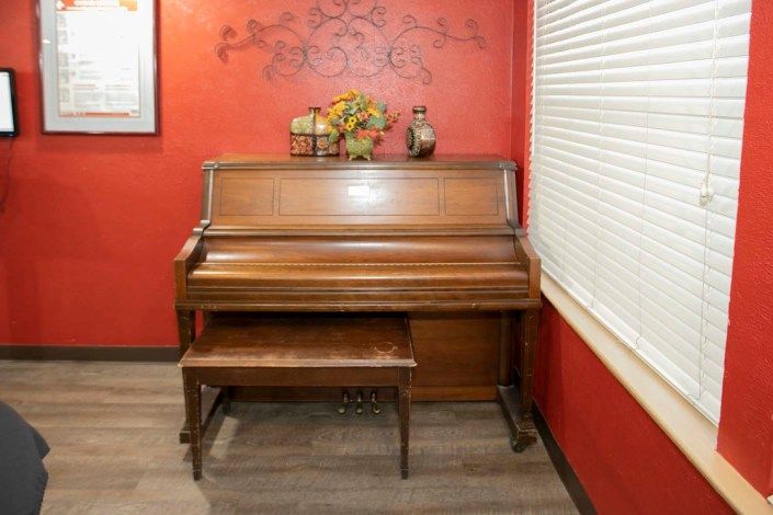 An upright wooden piano with a bench against a red wall adorned with decorative items.