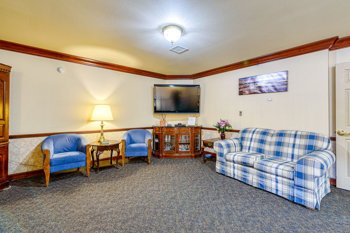 A cozy living room featuring a plaid sofa, two blue chairs, a television on the wall, and a lamp providing warm lighting.