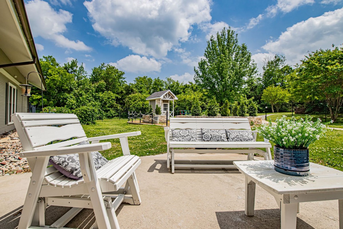 A sunny patio with white outdoor furniture overlooking a lush garden.