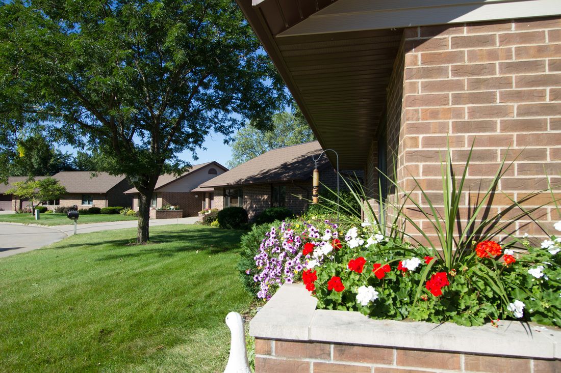 A suburban brick house with a colorful flower bed and a well-maintained green lawn on a sunny day.