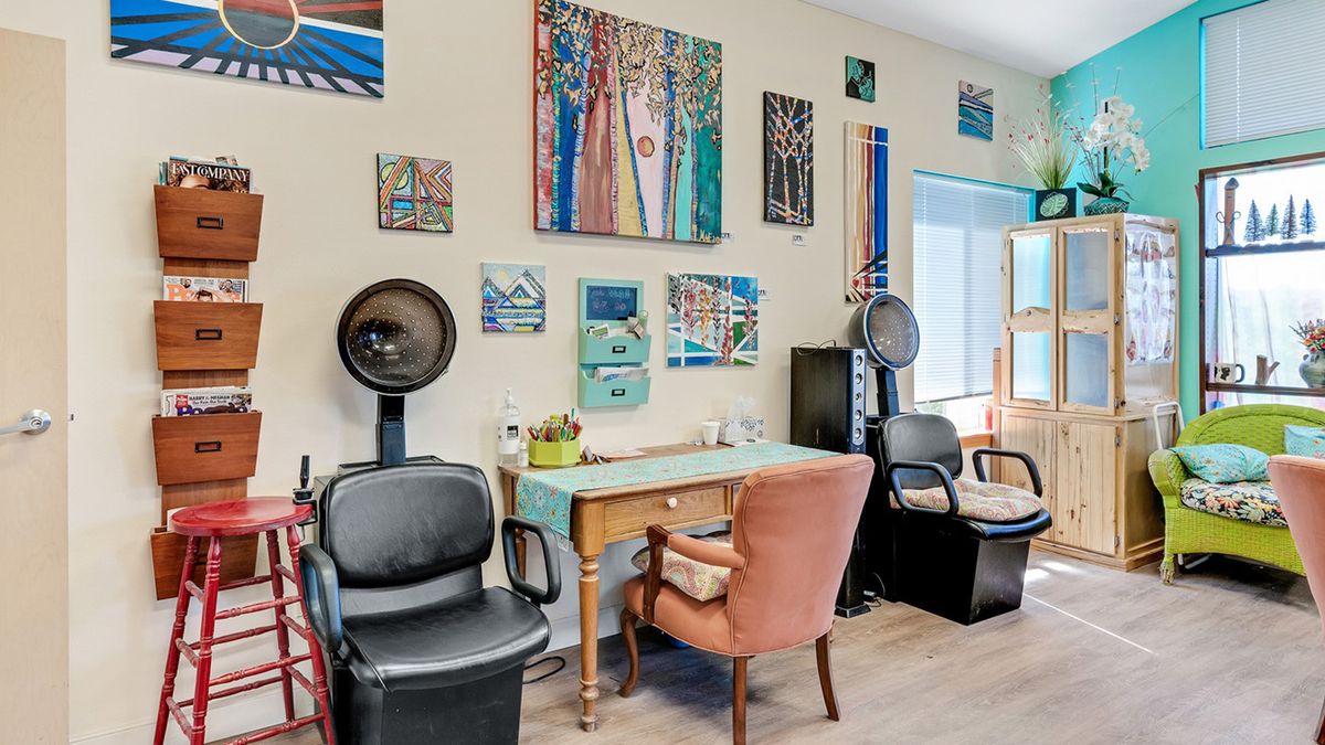 A cozy salon interior featuring hair dryer chairs, vibrant artwork on the walls, and a desk with an upholstered chair.