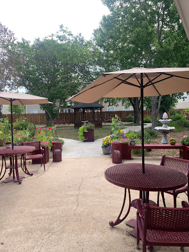 A serene outdoor garden space with tables under umbrellas, a pathway leading to the fountain, and lush greenery.