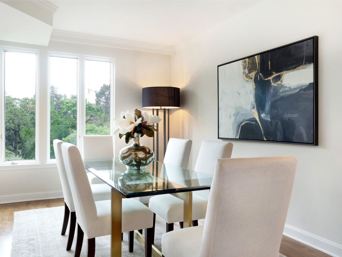 A modern dining room with a glass table and white chairs, adorned with abstract artwork and illuminated by natural light.