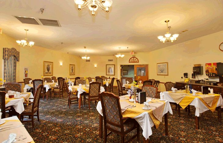 A warmly lit restaurant dining room with neatly arranged tables and chairs, elegant chandeliers, and a patterned carpet.