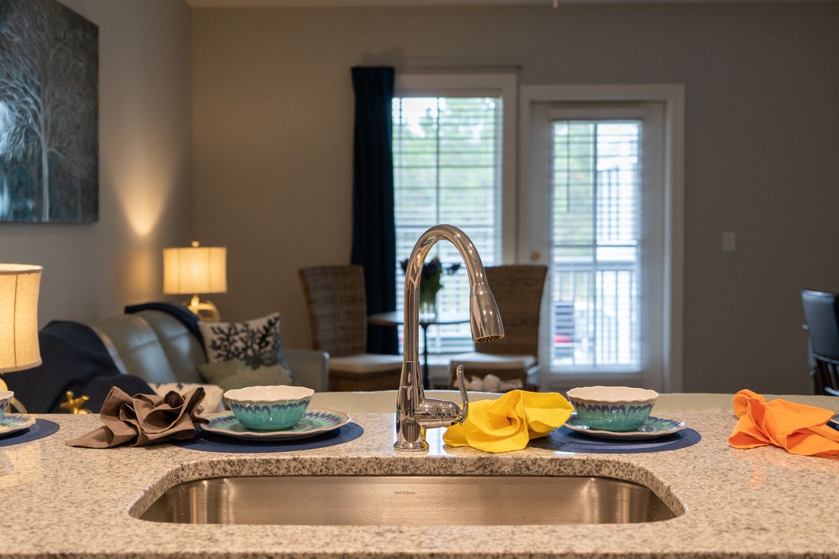 A modern kitchen with a view of the dining area through a window, featuring a sleek faucet and neatly arranged tableware on the countertop.