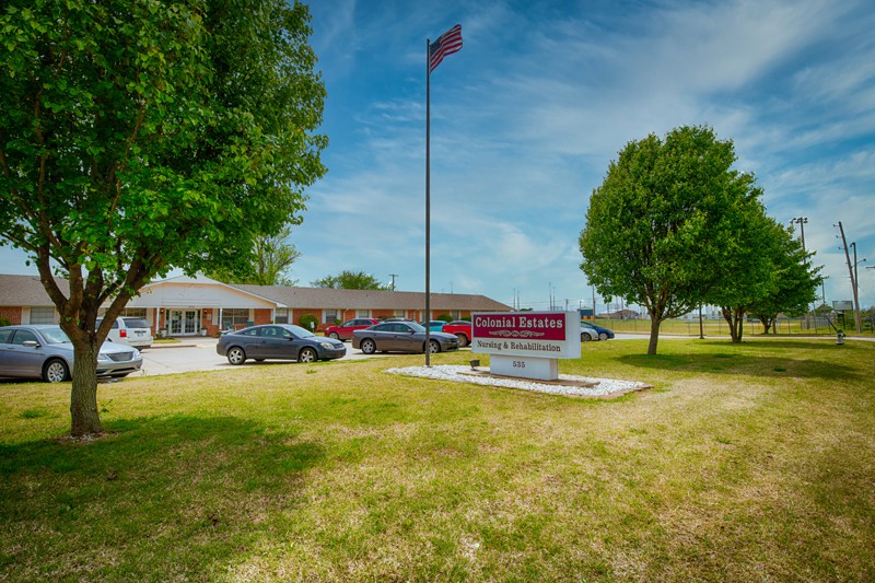 A school building with a parking lot, trees, and a flagpole on a sunny day.