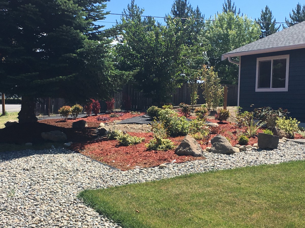 A beautifully landscaped garden featuring mulch, various plants, and rocks, next to a house.