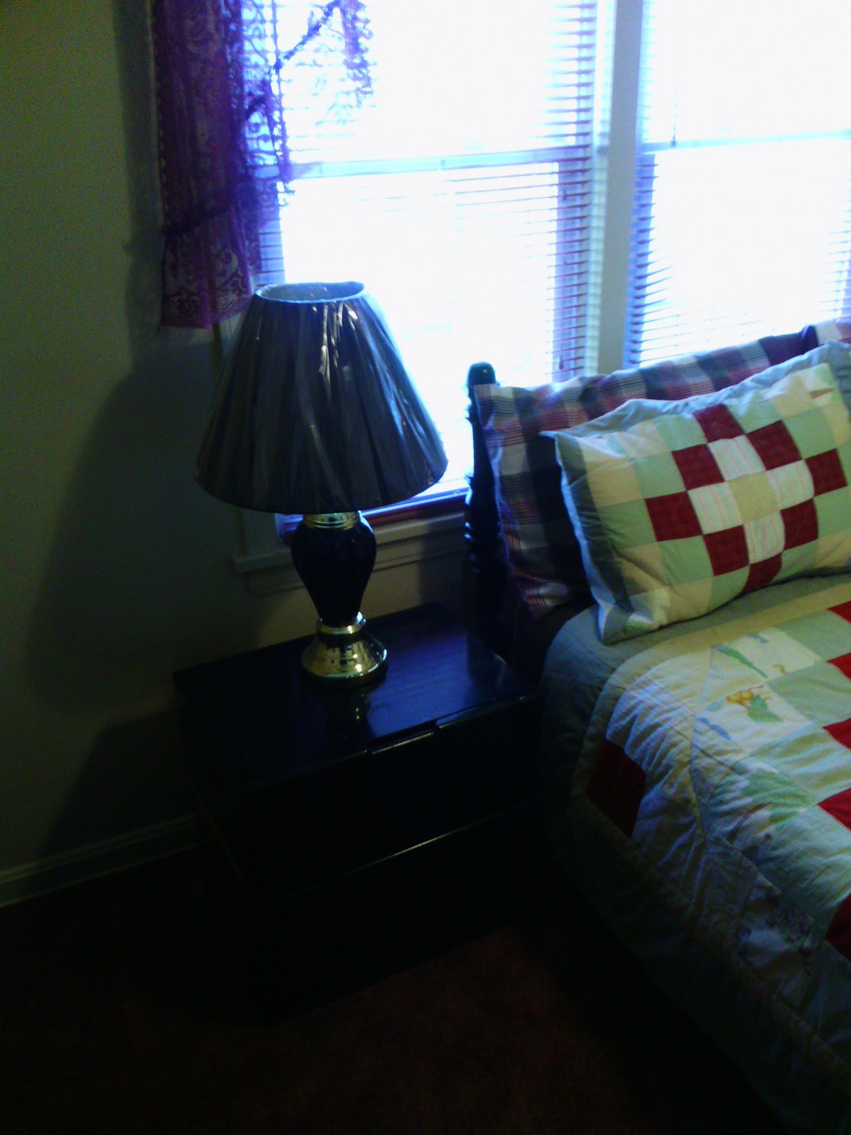 A cozy bedroom corner with a lamp on a bedside table next to a bed with a colorful quilt and pillow.