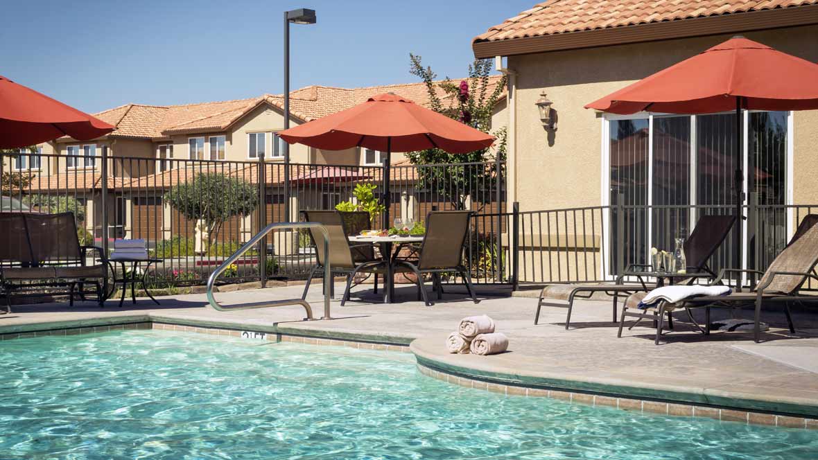 A sunny day by the swimming pool with patio furniture and umbrellas in a residential area.