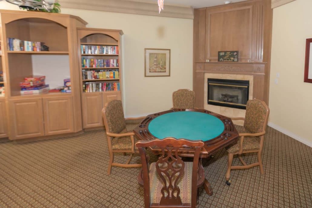 A cozy game room featuring a green poker table, surrounded by chairs, with a bookshelf and fireplace in the background.