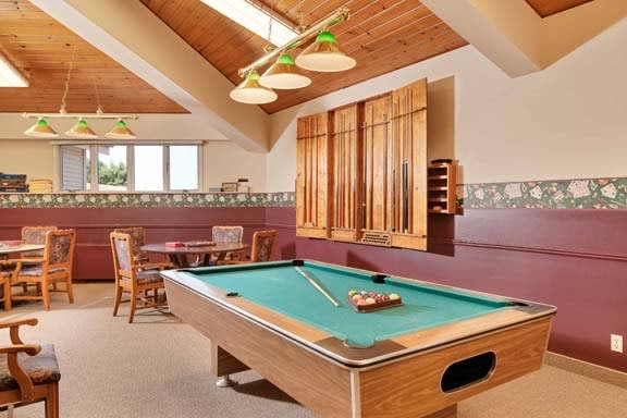 A cozy game room featuring a pool table under warm lighting, with wooden accents and additional seating in the background.