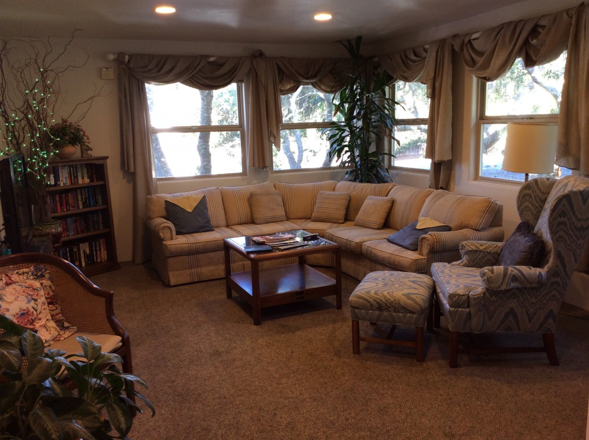 A cozy living room with a sectional sofa, coffee table, and multiple windows with curtains.