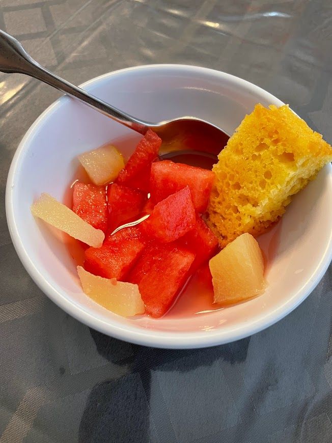 A bowl of fruit salad with watermelon and pineapple, accompanied by a piece of cake and a spoon.