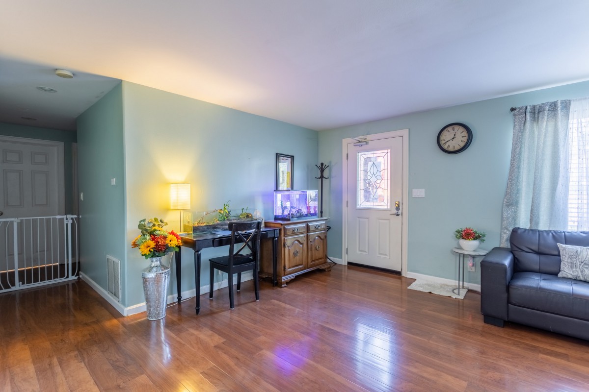 A cozy living room with hardwood flooring, featuring a table with a lamp and flowers, a fish tank, and a clock on the wall.