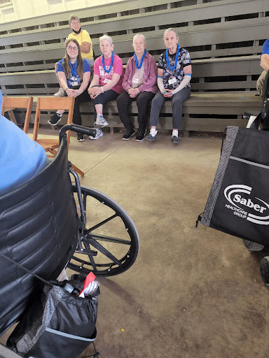 A group of individuals wearing medals seated together in an indoor setting, with a wheelchair in the foreground.