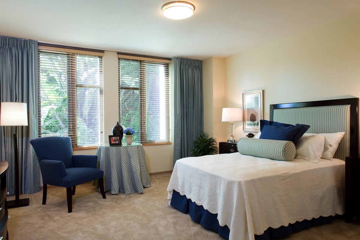 A cozy bedroom with a neatly made bed, a blue chair, and large windows covered with curtains.