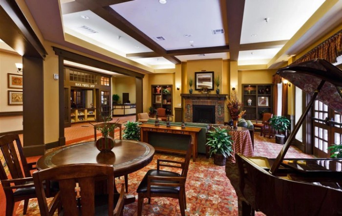 A cozy lounge area with a fireplace, surrounded by tables and chairs, adorned with plants and warm lighting.