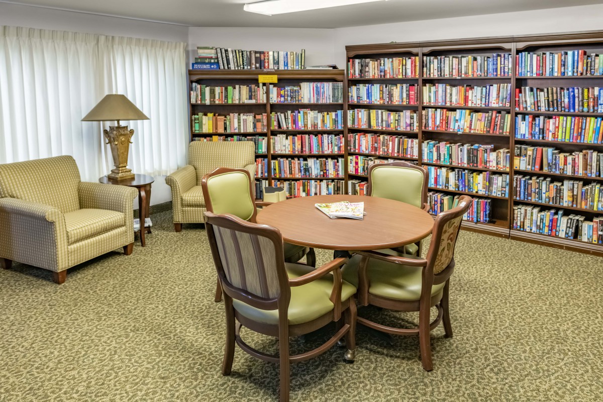 A cozy library reading area with bookshelves, a round table, and comfortable chairs.