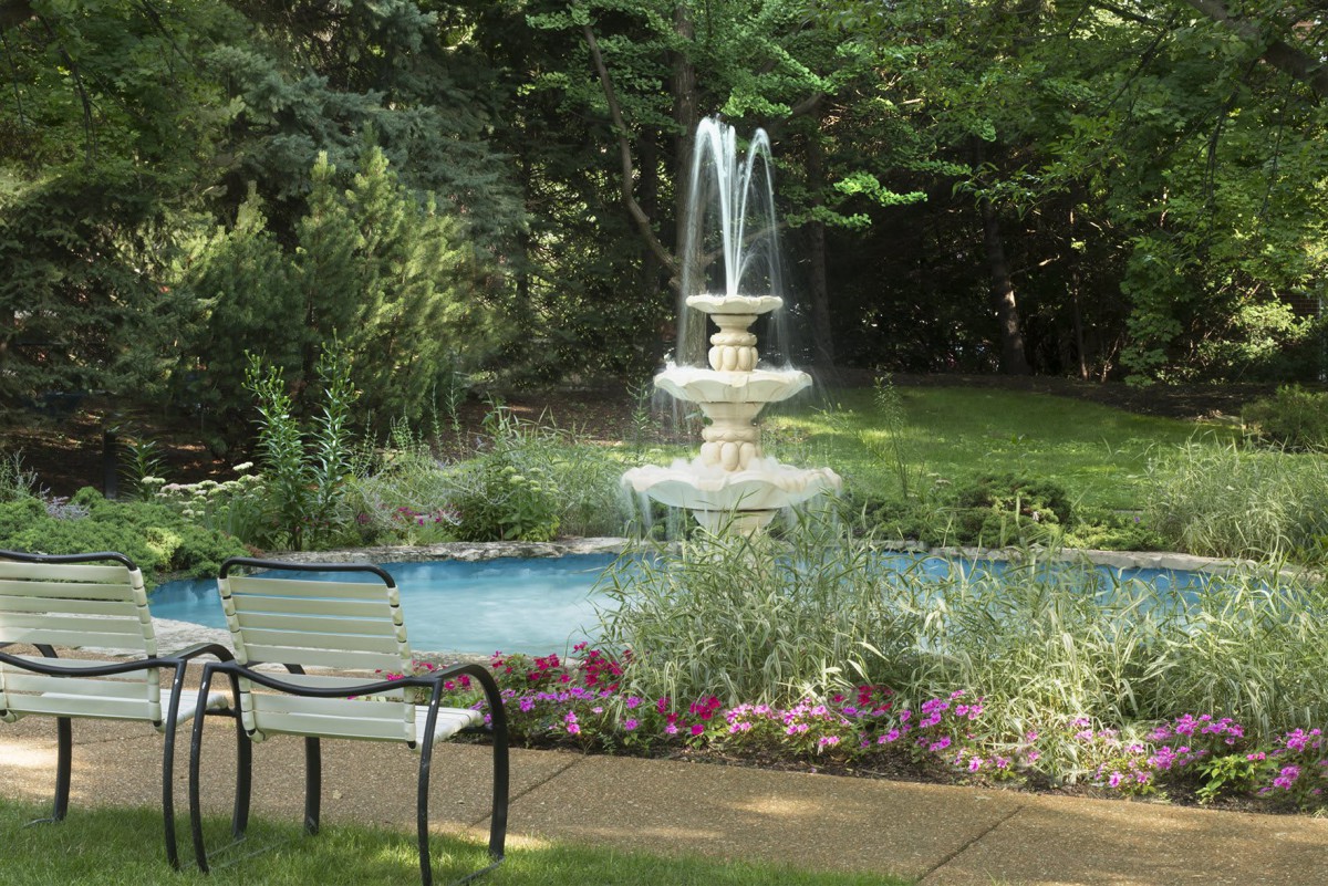 A serene garden scene with a water fountain and a nearby bench surrounded by lush greenery.
