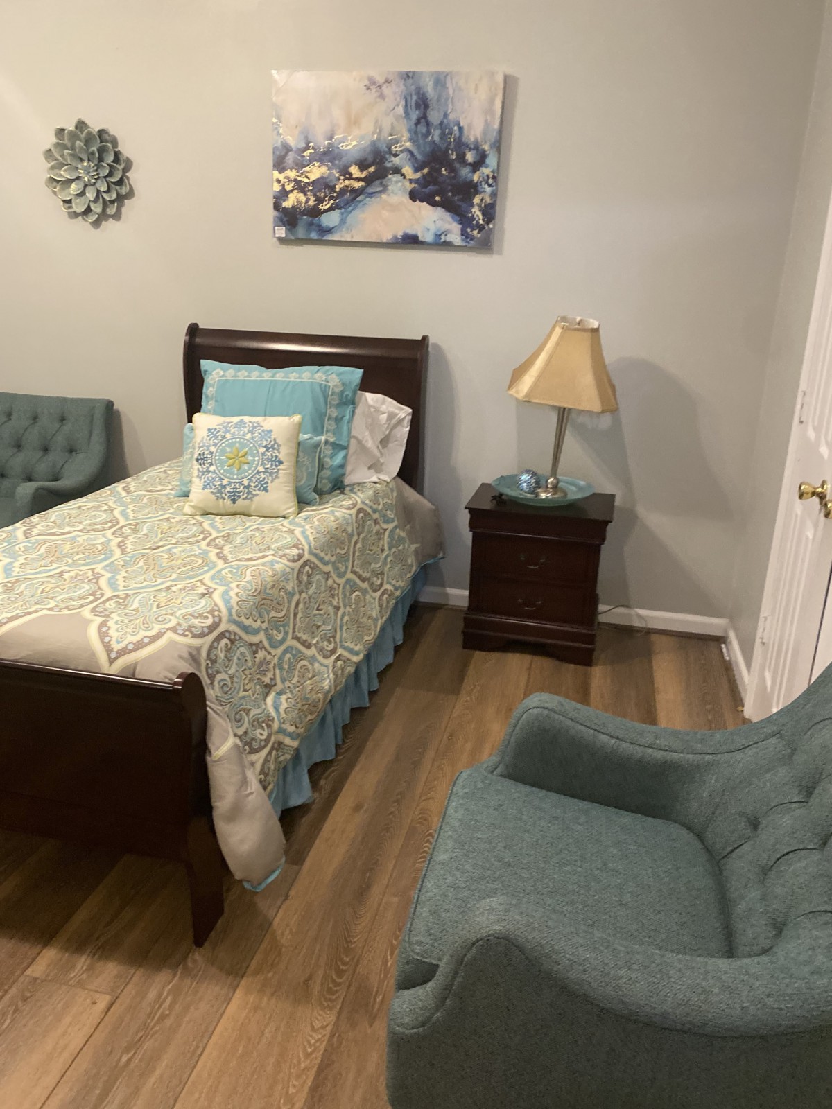 A cozy bedroom with a single bed, nightstand, lamp, and comfortable chair.