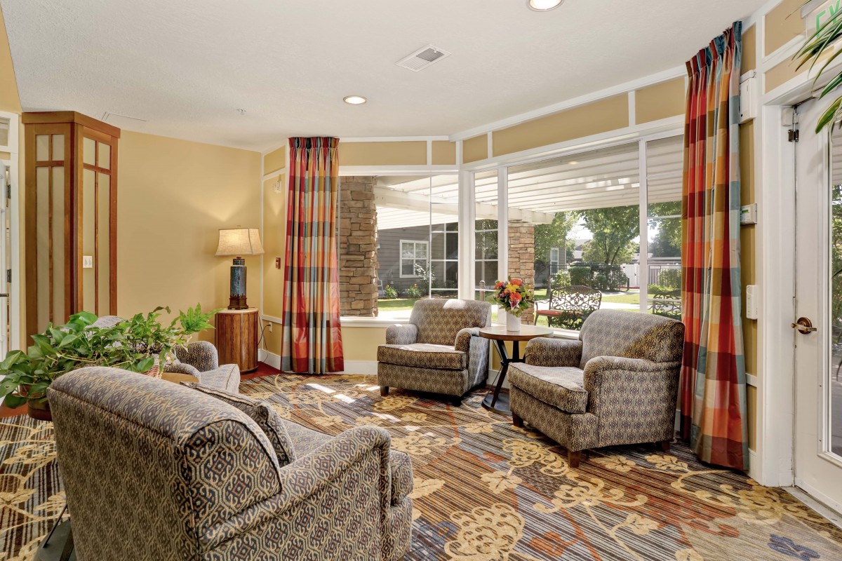 A cozy living room with patterned chairs, a lamp, and large windows letting in natural light.